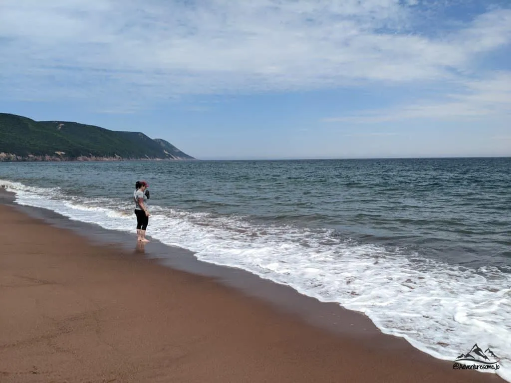 Touriste debout sur la plage du parc provincial Cabot Landing, les pieds dans le sable et les vagues frappant ses orteils.