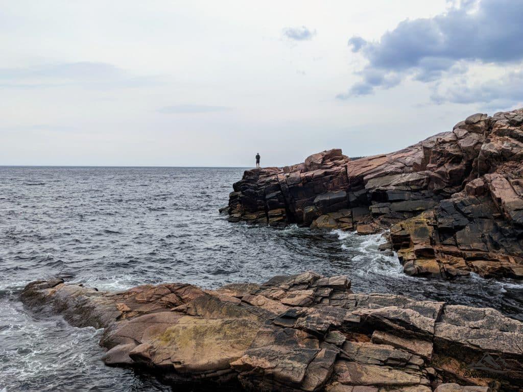 Hiker standing on top of the rocky edge of the water looking out to sea with waves hitting the rocky shore.