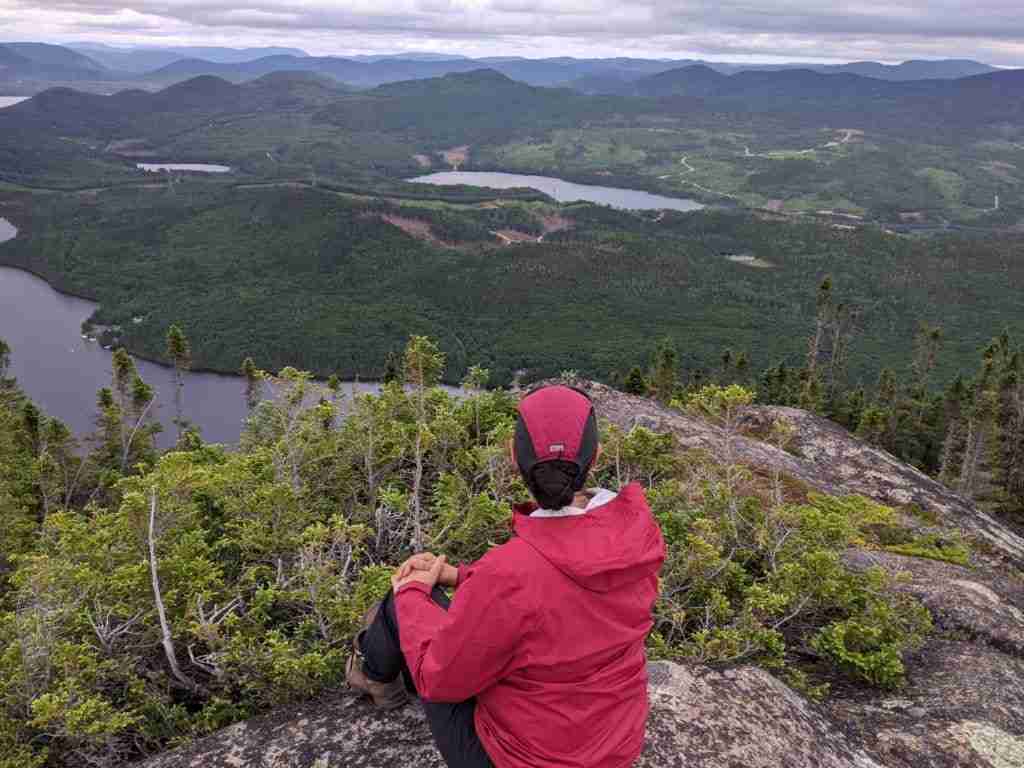 hiker sitting on a rock at the summit of mont Menaud looking at the surrounding hills and lakes
