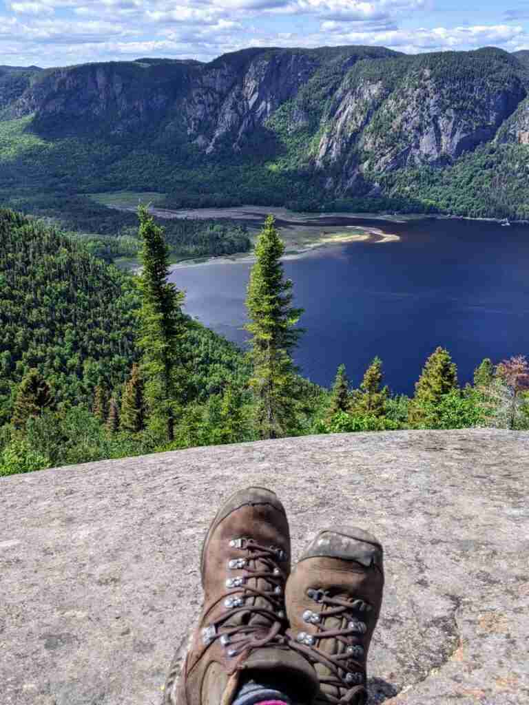 a hiker's boots showing crossed legs with the view of a river and the cliffs on the far shore