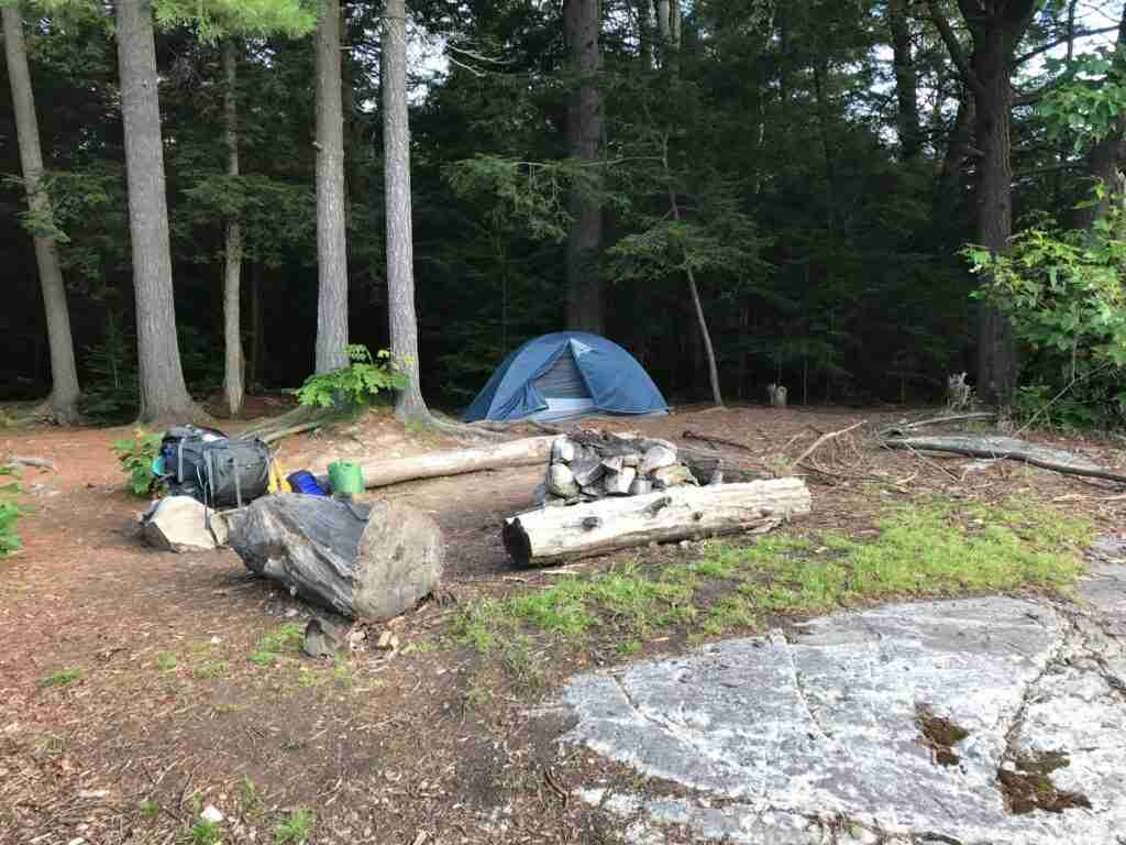 backcountry campsite on a backpacking trail in Ontario. The firering is made of rocks with two logs set on each side as seats and a tent in the backl