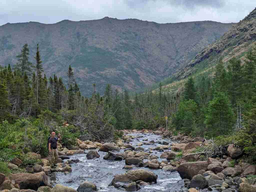 Randonneur sur le sentier traversant une rivière avec des rochers, bordée de conifères avec les montagnes en arrière-plan dans le parc national de la Gaspésie.