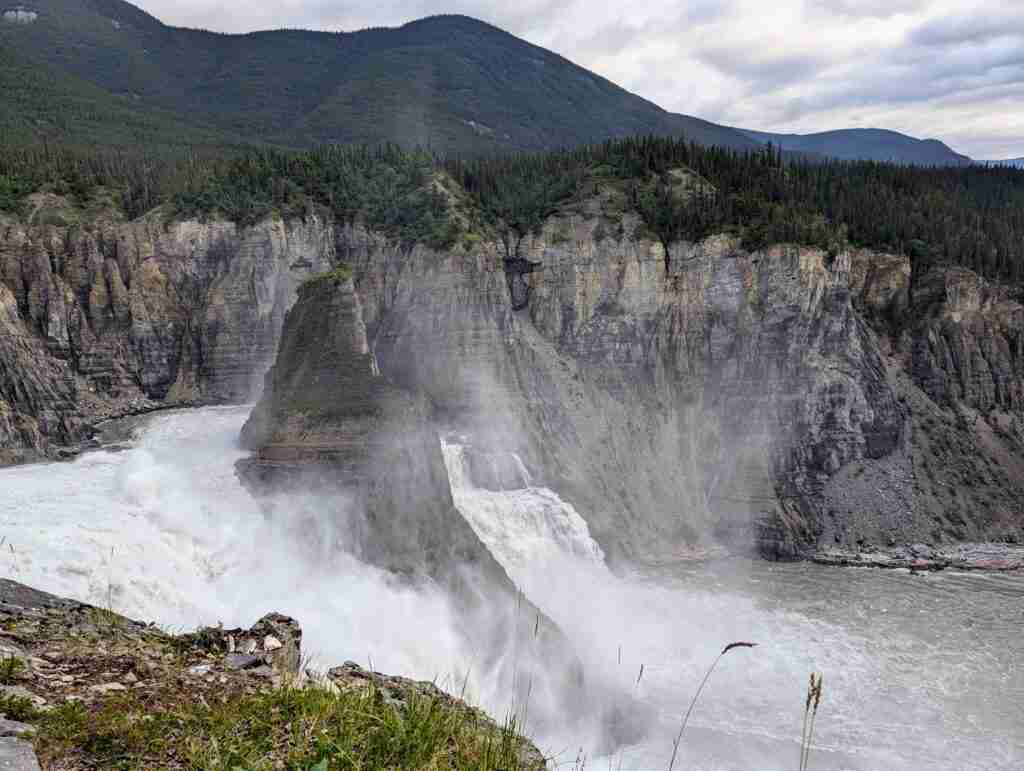 Virginia Falls with waters cascading down on each side of a tall rock pillar surrounded by towering cliffs