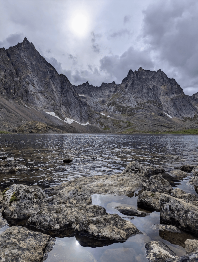 the rocky shore of Grizzly Lake with the grey jagged mountains on the far shore