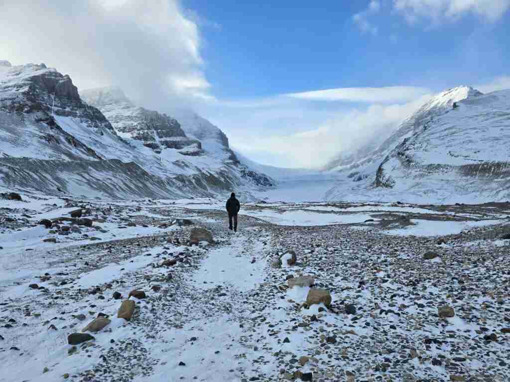 randonneur marchant sur un sentier rocheux et enneigé vers le glacier Athabasca avec de hautes montagnes enneigées de chaque côté dans le parc national Jasper