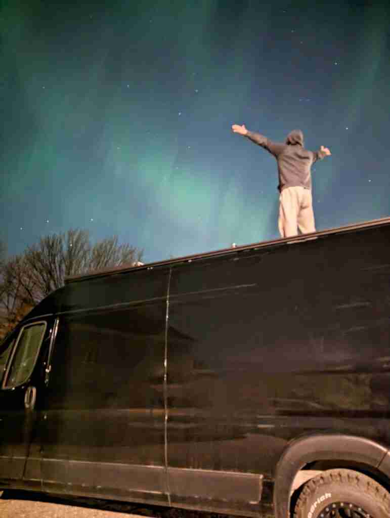 un homme debout sur le toit d'une camionnette, les bras tendus, regardant les aurores boréales vertes